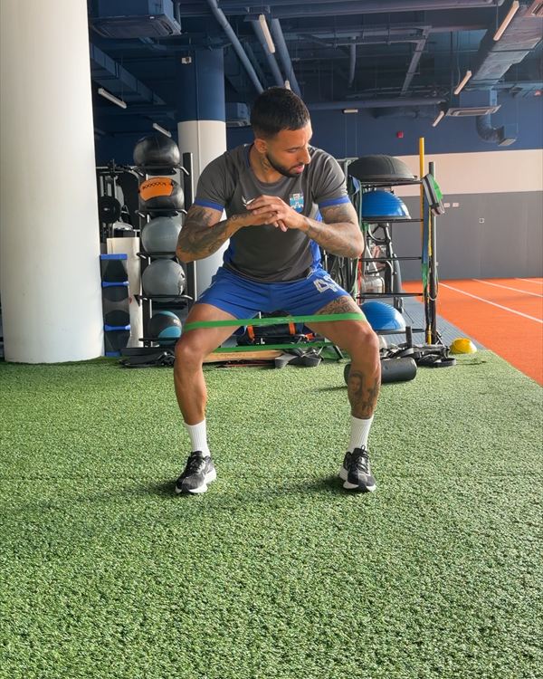 Renan Guedes faz um exercício de ativação com mini band elástica nos joelhos, em um espaço de treino.